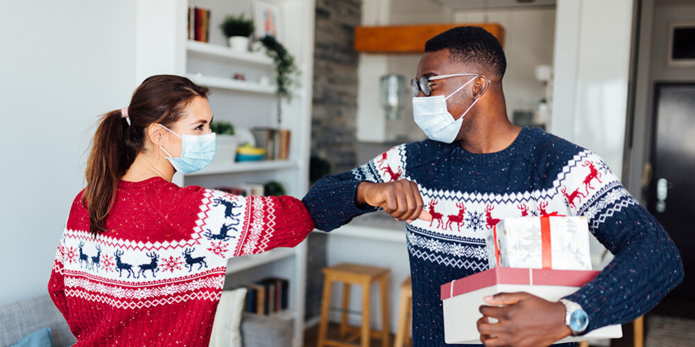 Young woman and man wearing holiday sweaters, elbow bumping and wearing masks