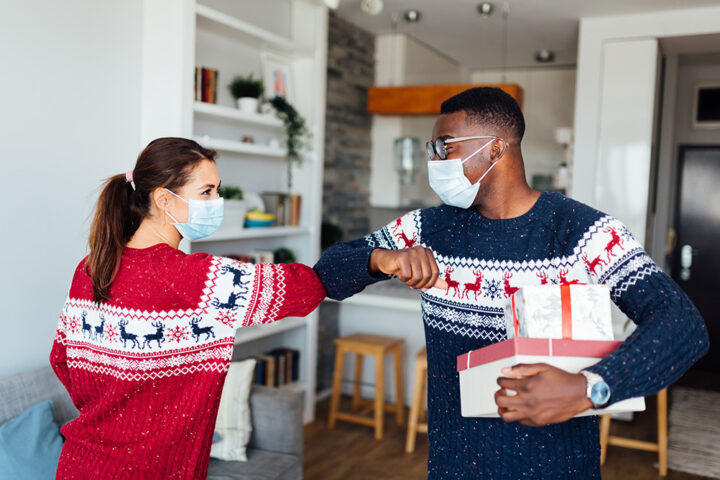 Young woman and man wearing holiday sweaters, elbow bumping and wearing masks